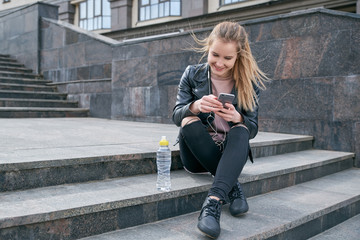 Young attractive smiling girl in a leather jacket and jeans sitting on the stairs with a smartphone...