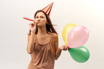 positive cheerful young girl in a party hat and with trumpet and baloons poses for a portrait with flowing long hair. Studio portrait on isolated white background. Holiday, birthday, achievement.