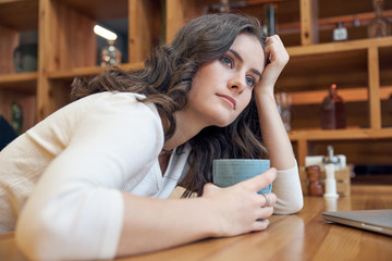 Beautiful Caucasian girl woman with long red hair sitting at a table with a mug of coffee or tea with a neutral expression looking into the distance about something thinking.