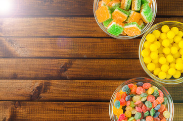 Brightly colored candies in glass cups on a wooden table, top view, free space for text