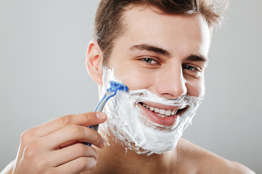 Photo of masculine brunette guy with dark short hair shaving his face with razor and gel or cream being satisfied over grey background close up