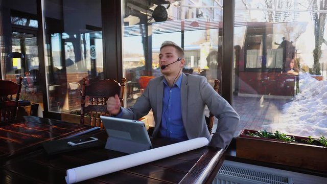Young draftsman talking with client by tablet, headset phones and microphone at cafe.