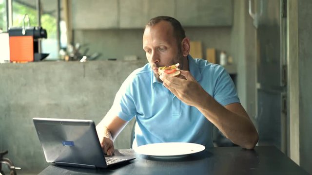 Young man with laptop eating breakfast sitting by table in kitchen at home
