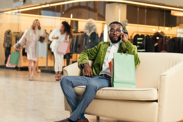 stylish african american man talking by smartphone while sitting with shopping bags in mall