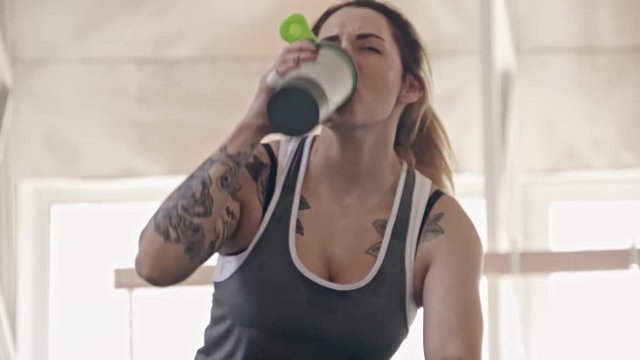 Tired woman with muscular tattooed arms resting in gym and drinking water after workout