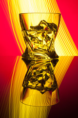 Cocktail whiskey a glass with pieces ice of party reflection a concept of hourglasses yellow light effects on red background.