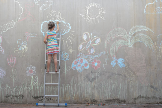 Girl standing on ladder drawing colorful pictures with chalk on a concrete wall