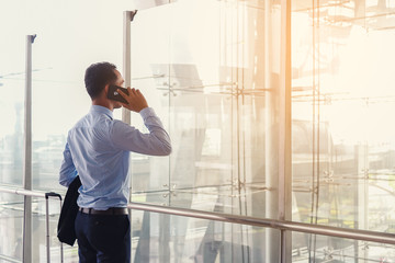 Back side of handsome business traveler using his smart phone for business connection at airport terminal. Businessman standing with luggage and his suit, using smart phone before plane take off.