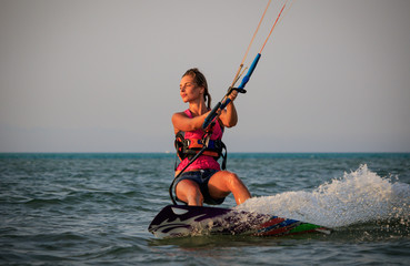 Fototapeta premium Kite surfing girl in sexy swimsuit with kite in sky on kiteboard in the blue sea riding waves saying hi. Recreational activity, water sports, action, hobby and fun in summer time. Kiteboarding sunset