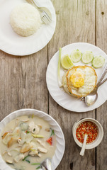 authentic Thai dinner background: Tom kha gai spicy soup, plain rice and pad kra pao with fried egg on top and homemade chilli sauce on wooden table at local cafe. Asian food concept. Text space