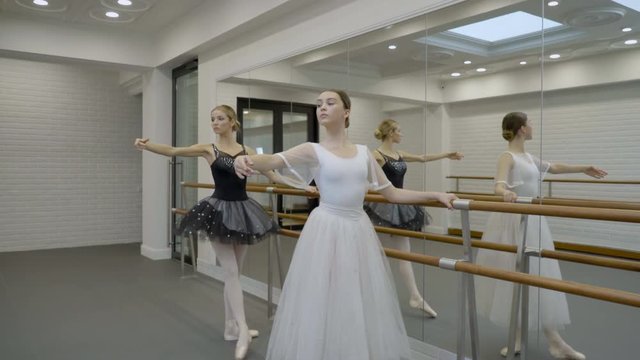 The two prima ballerinas are exercising while standing near the barre in big ballet class with mirror. The dancers are doing inclines and passe par terre