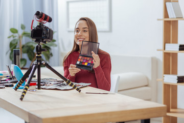 Shadow palette. Cheerful enthusiastic young blogger feeling glad while smiling to the camera and showing a wonderful shadow palette
