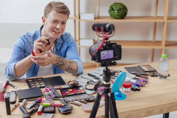 Blog recording. Young smart positive man feeling good while being surrounded by various cosmetics and recording a video blog about the usage of lipsticks