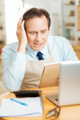 Incorrect calculations. Senior occupied disappointed man sitting by the table opposite the laptop holding right hand near head and overlooking the notebook.