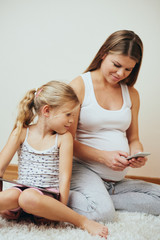 Pregnant woman holding smartphone sitting in the room with her daughter.