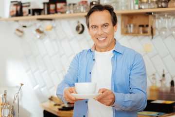 Take it please. Handsome pleasant polite man standing in the kitchen smiling and giving a cup of coffee.