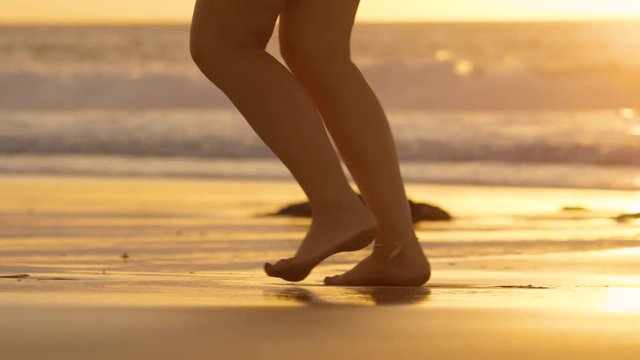 SLOW MOTION, LENS FLARE: Unrecognizable woman running barefoot on wet sand on a summer evening. Active female with ankle bracelet jogging on a sunny tropical beach as ocean waves reach wet sandy coast