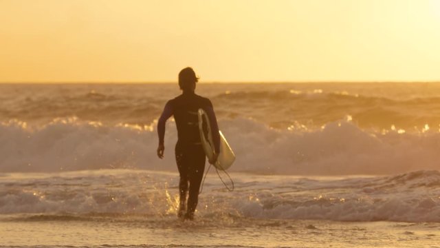 SLOW MOTION: Young surfer running into water to surf breaking waves at golden sunrise. Carefree male surfboarder jogging into ocean to ride waves on his cool surfboard in the beautiful summer sunset.