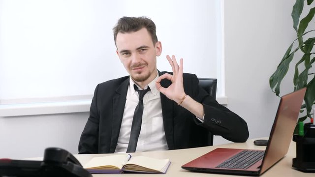 Young businessman in suit sitting in office and showing ok sign. 60 fps