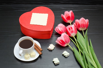 Red box in heart shaped, pink tulips, gray sheet and a coffee mug. Black background