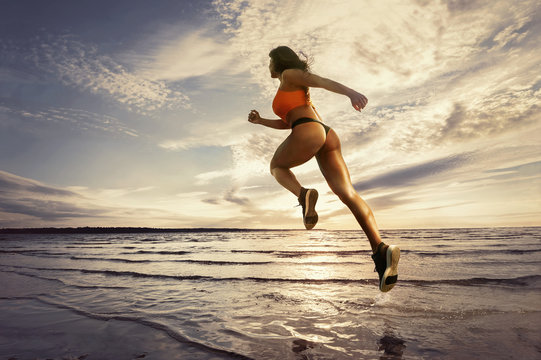 Young athletic woman running and jumping in water during workout on the beach at sunset