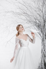 Fototapeta na wymiar A young blonde bride in white wedding dress on a background of white walls and white tree in the background holds a white dove