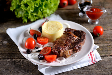Beef steak with mashed potatoes and salad of cherry tomatoes, dried olives and white onions.