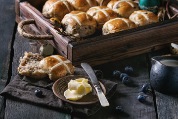 Hot cross buns in wooden tray served with butter, knife, blueberries, easter eggs, birch branch, jug of cream on textile napkin over old texture wood background. Dark rustic style. Easter baking.