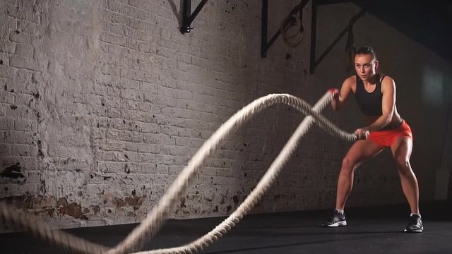 Concept: power, strength, healthy lifestyle, sport. Powerful attractive muscular woman Cross Fit trainer do battle workout with ropes at the gym. Slow motion