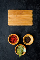 Obraz na płótnie Canvas Mock up for menu or recipe. Wooden cutting board near bowls with goji, bay leaf, pepper on black background top view