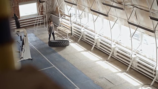 High Angle View Of Strong Female Athlete Flipping Large Tractor Tire During Cross-training Workout In Dusty Old Gym