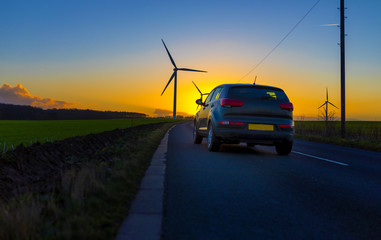 Car driving down country road towards wind turbines in a field in the UK at sunset or sunrise on  a clear winter day