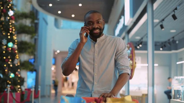 young african american man walking in a shopping center talking on a phone smiling feeling happy near a Christmas tree smartphone black buy close up mall shop store attractive slow motion