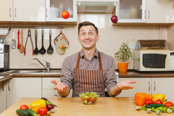 Fun crazy caucasian young man in apron, brown shirt sitting at table, throwing up onion and tomato,...
