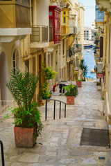 Fototapeta na wymiar Medieval town narrow street with stairs in Valletta on the island of Malta. Colorful red, yellow, blue balconies and doors and green plants near traditional style stone buildings. Sea side view.