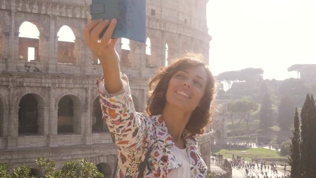 Beautiful young woman in colorful fashion dress alone on hill with bike taking selfies with colosseum in Rome with smartphone at sunset attractive elegant girl