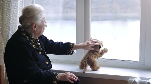 a lonely old woman looking out the window with a soft toy on the windowsill