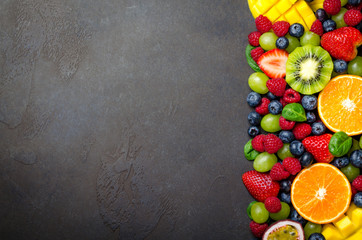 Various fresh fruits: strawberry, raspberry, blueberry, tangerine, grape, mango, spinach on a dark black stone background. Copy space, top view, horizontal image