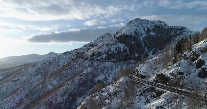 Backward aerial top view over car travelling on road in Winter snow mountain near forest woods.Snowy mountains street path establisher.Sunset or sunrise sun backlit.4k drone flight  establishing shot