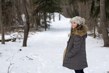 Beautiful brunette young woman in winter scene - snow covered park wearing winter clothes - parka with knit cap - standing on snow covered trail looking back over shoulder