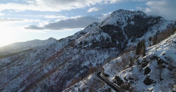 Backward aerial top view along road on white snow mountain in winter.Forest woods.Snowy mountains street path establisher.Sunset or sunrise sun backlit.4k drone flight straight-down establishing shot