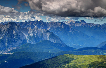 Jagged sharp ridges and vertical rock faces of Carnic Alps and Prealps / Friuli Dolomites and white cumulus clouds above forests and Piave river valley in blue summer mist, Belluno Veneto Italy Europe