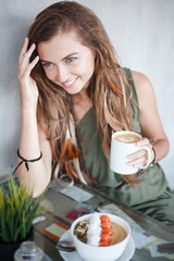 Beautiful smiling girl with cup of coffee and muesli bowl is sitting in a cafe