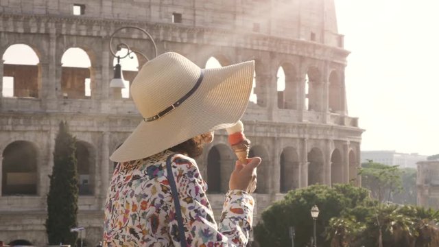 Beautiful young woman in colorful fashion dress eating ice cream in front of colosseum in Rome at sunset attractive girl with elegant straw hat