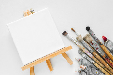 Canvas on easel, paint tubes and bundle of brushes for painting on white canvas background. Top view. Flat lay.