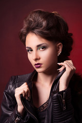 Fototapeta premium Brunette girl with bright makeup and stylish hairstyle holding hands on the collar of a leather jacket on a dark red background.