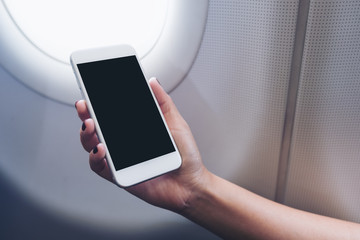 Mockup image of a hand holding and looking at white smart phone with blank desktop screen next to an airplane window with clouds and sky background