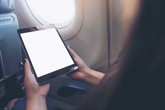 Mockup image of a woman holding and looking at black tablet pc with blank white desktop screen next to an airplane window with clouds and sky background