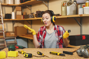 Perplexed young woman in plaid shirt gray T-shirt noise insulated headphones yellow gloves working in carpentry workshop at wooden table place with piece of wood, different tools, power drill, wire.
