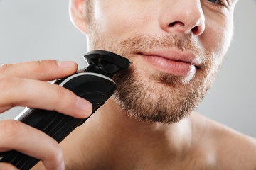 Close up shot of handsome man smiling while shaving his face with electric shaver making morning...